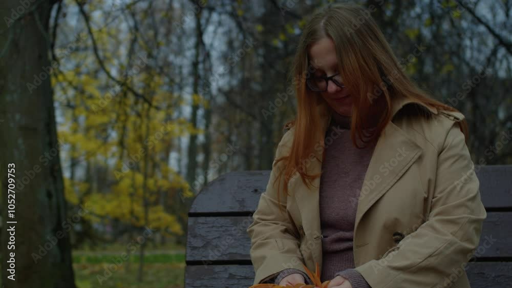 A woman in glasses, a warm sweater and coat sits on a bench in an autumn park, folds something from leaves and laughs
A woman in glasses, a warm sweater and coat sits on a bench in an autumn park, folds something from leaves and laughs