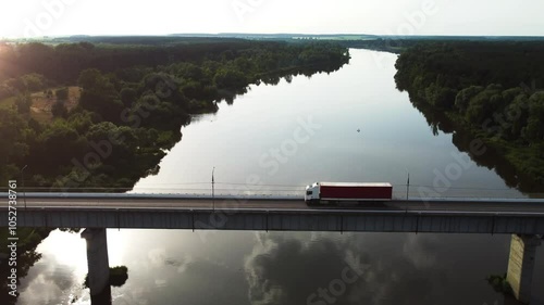 White truck with a red semi-trailer drives on a highway bridge over a river during sunset. Lorry is hauling container trailer. Aerial drone view. Concept of cargo delivery, logistics, transportation. 