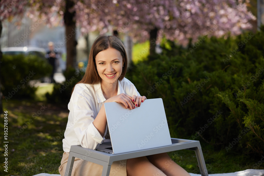 smiling woman, outdoor tech, scenic view. Smiling woman sits outdoors in park, working on laptop under blooming trees, enjoying peaceful, natural setting. smiling woman, outdoor tech, scenic view. Smiling woman sits outdoors in park, working on laptop under blooming trees, enjoying peaceful, natural setting.