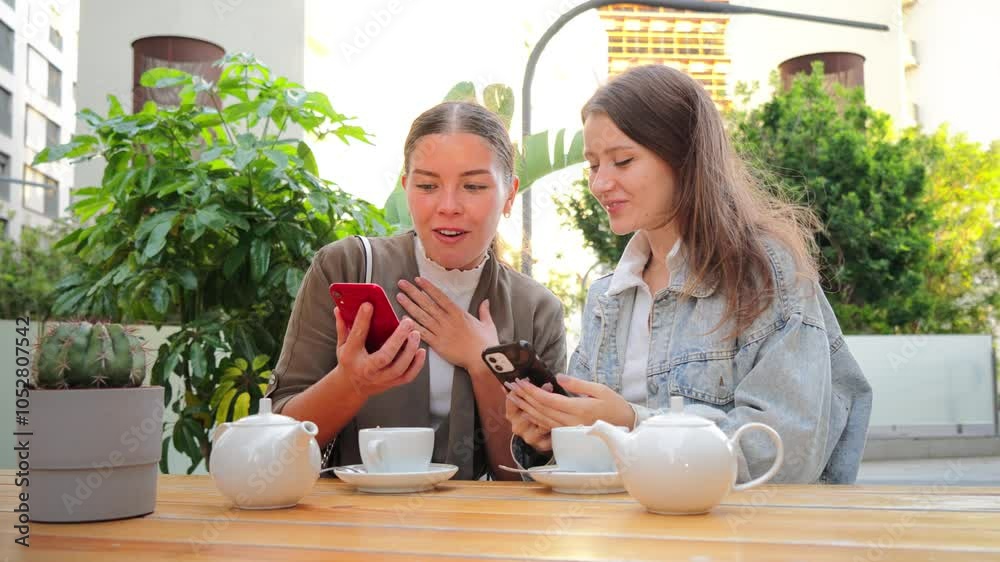 Young women enjoying a tea together while watching and sharing on a cellphone, smiling and relaxing outdoors, connecting and bonding over social media in a casual, friendly coffeeshop terrace