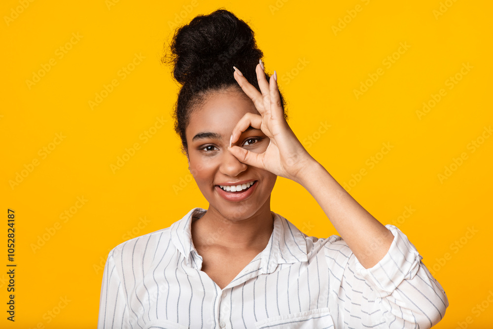 Smiling Black Woman Looking Through Ok Sign, Covering One Eye With Okay Gesture And Looking At Camera, Yellow Background With Free Space Smiling Black Woman Looking Through Ok Sign, Covering One Eye With Okay Gesture And Looking At Camera, Yellow Background With Free Space
