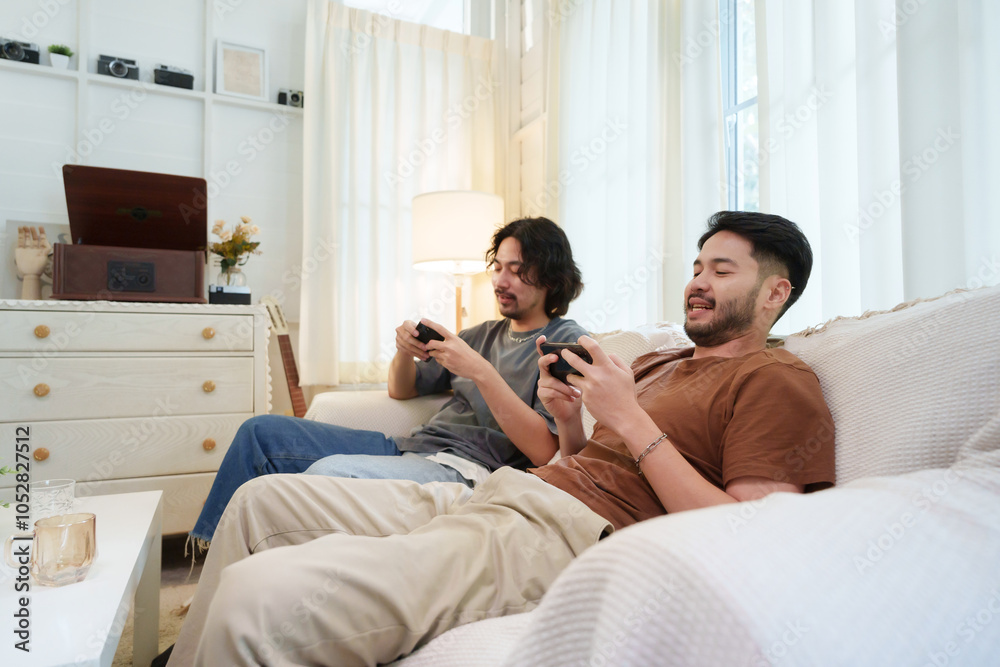 Two Asian men sitting on a sofa, focused on mobile gaming on smartphones, Two friends enjoying a relaxed moment at home. Two Asian men sitting on a sofa, focused on mobile gaming on smartphones, Two friends enjoying a relaxed moment at home.