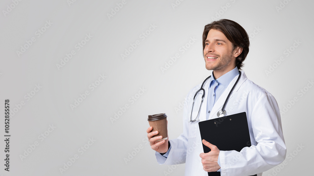 Happy young doctor holding clipboard with medical info and having coffee break over light studio background, free space Happy young doctor holding clipboard with medical info and having coffee break over light studio background, free space