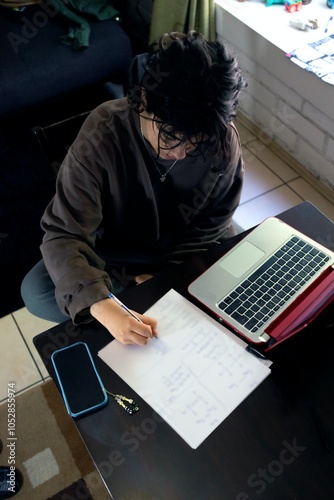 teenage girl writes in her notebook while studying on laptop computer