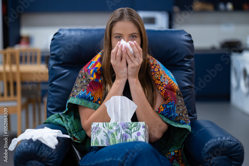 Sick young female with an illness, flu or cold sits at home using a tissue. Keeping warm under a blanket on the couch. Fighting symptoms of respiratory infection. Using tissues for a runny nose. photo