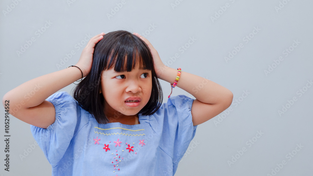 Frustrated cute Asian child girl wearing blue dress holding on head being depressed regretting what he did having headache, looking stressful.