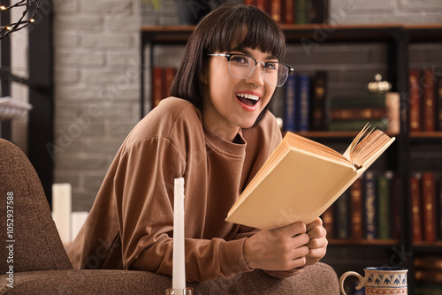 Smiling young woman with glasses reading a book in cozy home library, happy f...