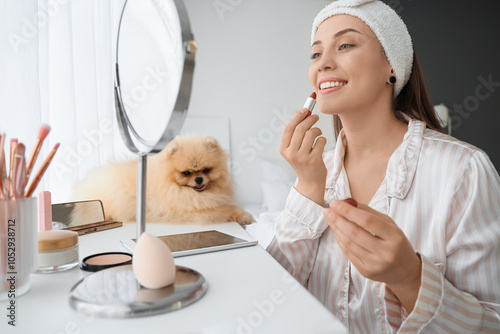 Young woman with cute Pomeranian dog applying lipstick in bedroom