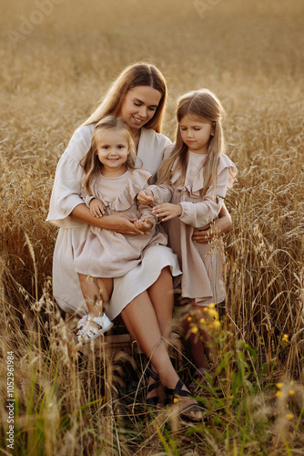 Young blonde woman with two daughters in beige dresses in a wheat field during sunset. Happy family walking.