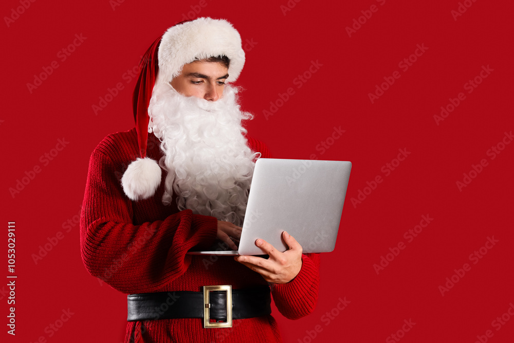 Naklejka premium Young Santa Claus using laptop on red background