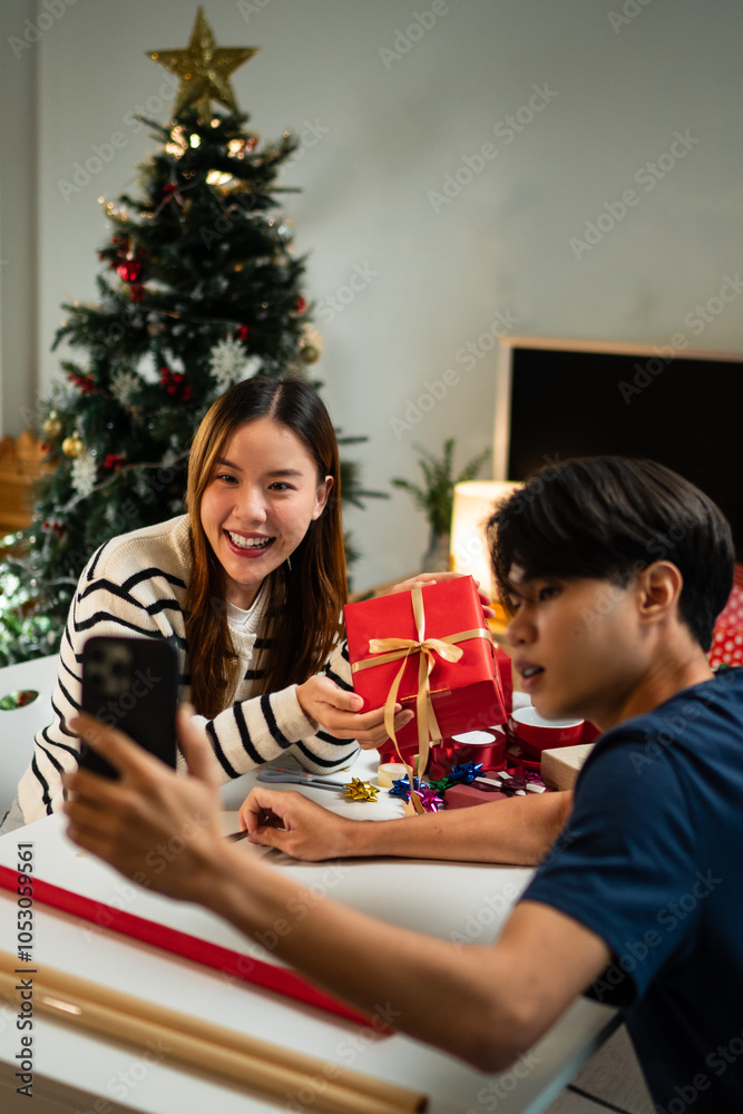 A young man and woman are sitting together at a table, smiling and taking a selfie during a festive moment. The table is filled with wrapped gifts, and a Christmas tree in the background. A young man and woman are sitting together at a table, smiling and taking a selfie during a festive moment. The table is filled with wrapped gifts, and a Christmas tree in the background.