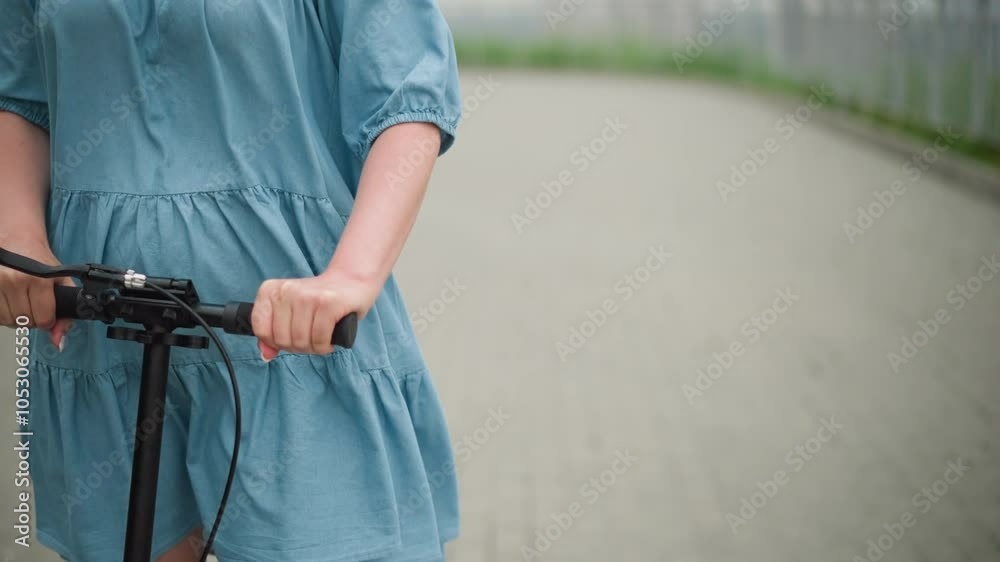 A mid-body shot of a woman riding a scooter on a pathway, her blue dress flutters in the wind as she grips the handlebars with both hands, with a blur background