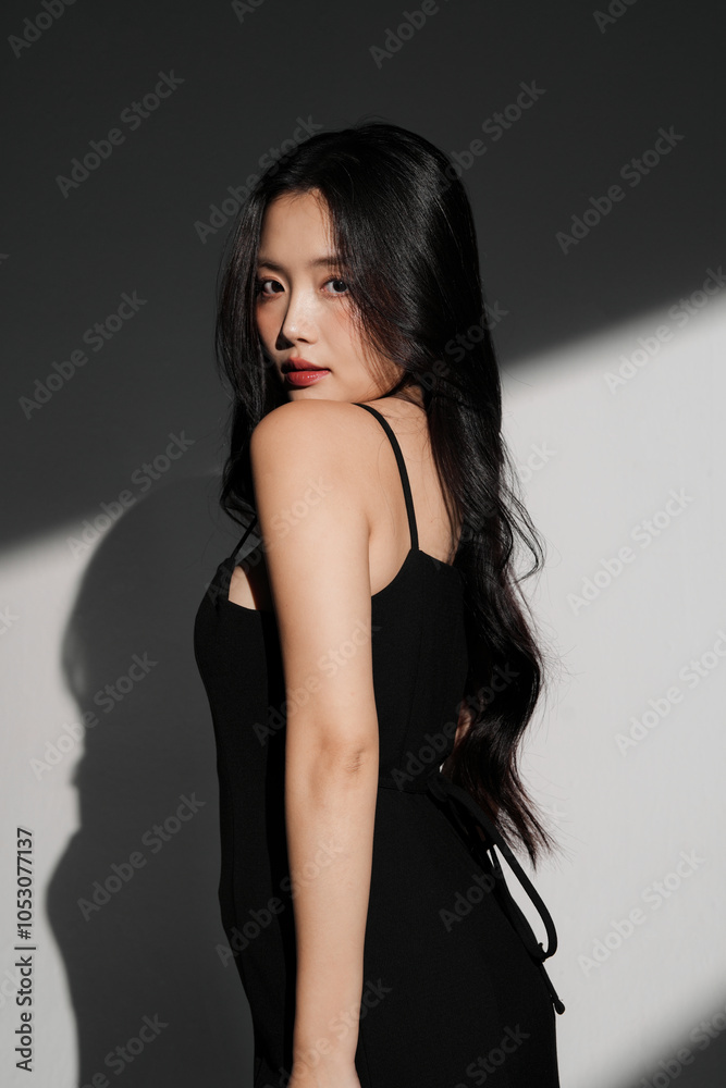 Fototapeta premium 光と影の背景で撮影をした黒いドレスを着た若い女性のシックなポートレート
