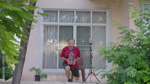 An Asian man in sports clothing performs lunges with a water container in front of his house, recording the session on his smartphone to track progress and share on social media