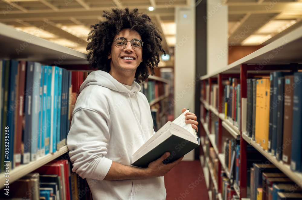 Dark-haired young guy standing near the bookshelves and reading a book