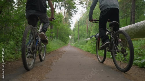 Two brothers riding bicycles along a paved untarred path surrounded by lush green trees, one brother stands while cycling, and a log of wood lies along the side of the road photo