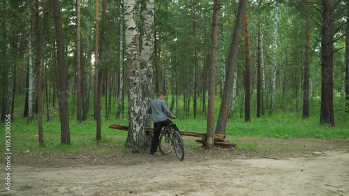 Back view of cyclist riding on an untarred road surrounded by tall birch trees in a forest, the cyclist is wearing a grey jacket, parked close to a tree, with another rider following from behind photo