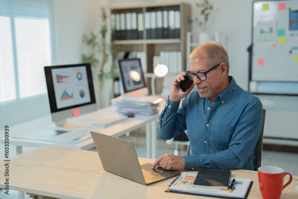 Senior businessman multitasks on laptop and phone at cluttered desk in bright office, focused and professional Senior businessman multitasks on laptop and phone at cluttered desk in bright office, focused and professional