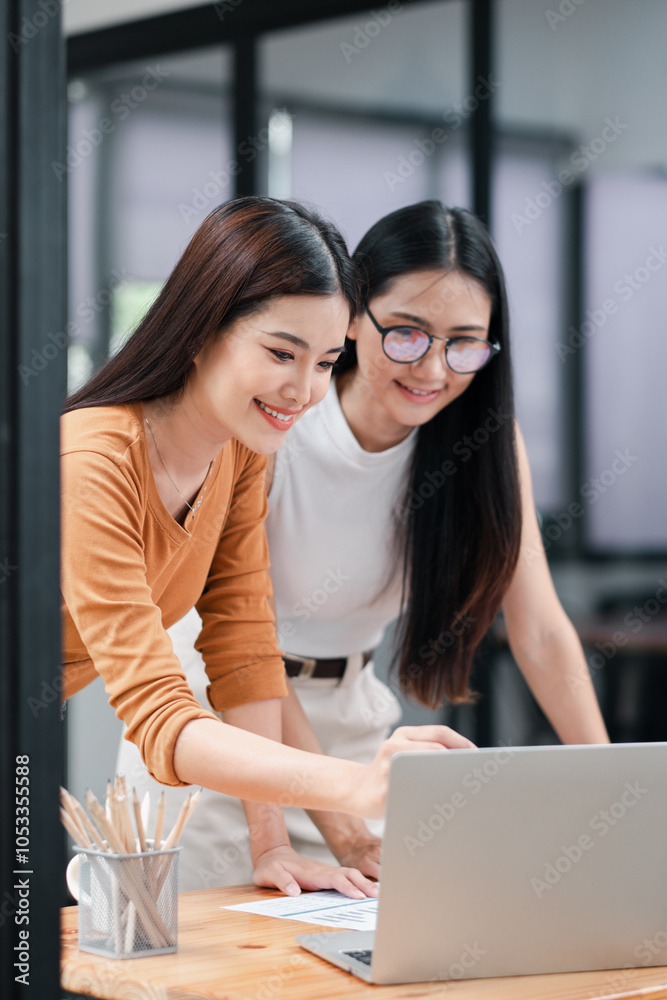 Two young women working together at a desk with a laptop, smiling and collaborating in a modern office setting. Two young women working together at a desk with a laptop, smiling and collaborating in a modern office setting.