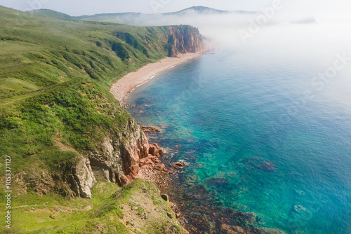 The stunning coastline displays rugged cliffs meeting a clear blue sea, with a tranquil sandy beach nestled amid lush greenery, enveloped in morning mist.Vladivostok russian far East