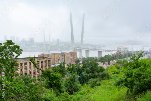 A bridge towers above the bay in Vladivostok, shrouded in fog. Cars navigate the roadway as grey clouds obscure the skyline and landscape. Vladivostok, Russia, 08.07.2022. photo