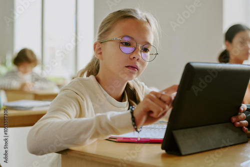 Young girl wearing glasses sitting at wooden desk using tablet in classroom surrounded by fellow students in bright learning environment photo