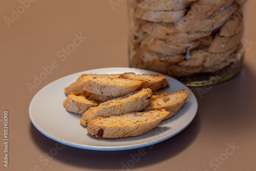 Cantucci almond cookies arranged on a white plate with a jar full of more cookies in the background highlight their crunchy texture and nutty flavor typical of traditional Italian biscotti photo