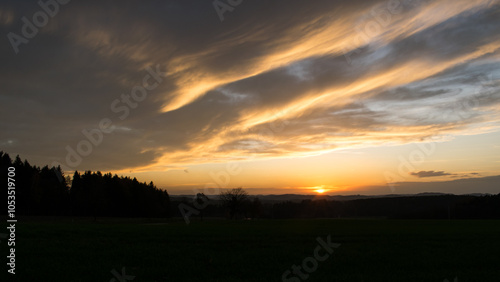 An autumn landscape at sunset, featuring a dramatic sky filled with vibrant colors, dominated by beautifully shaped clouds that are illuminated by the warm hues of the setting sun.