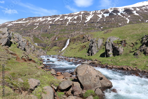 panoramic view of an icelandic landscape with a wild mountain river in a rocky terrain with numerous waterfalls coming down the mountain cliff in the distance that is stil partially covered with snow photo