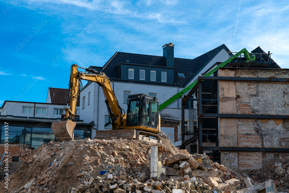 Obraz premium An excavator operates at a construction site, clearing debris from a demolished structure while a green crane is positioned nearby under a blue sky.