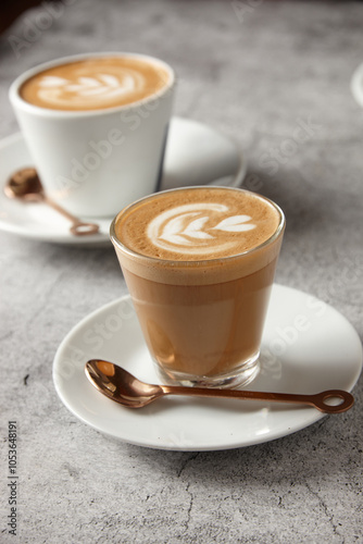 two cups of coffee with latte art on top on a white saucer with copper-coloured spoon