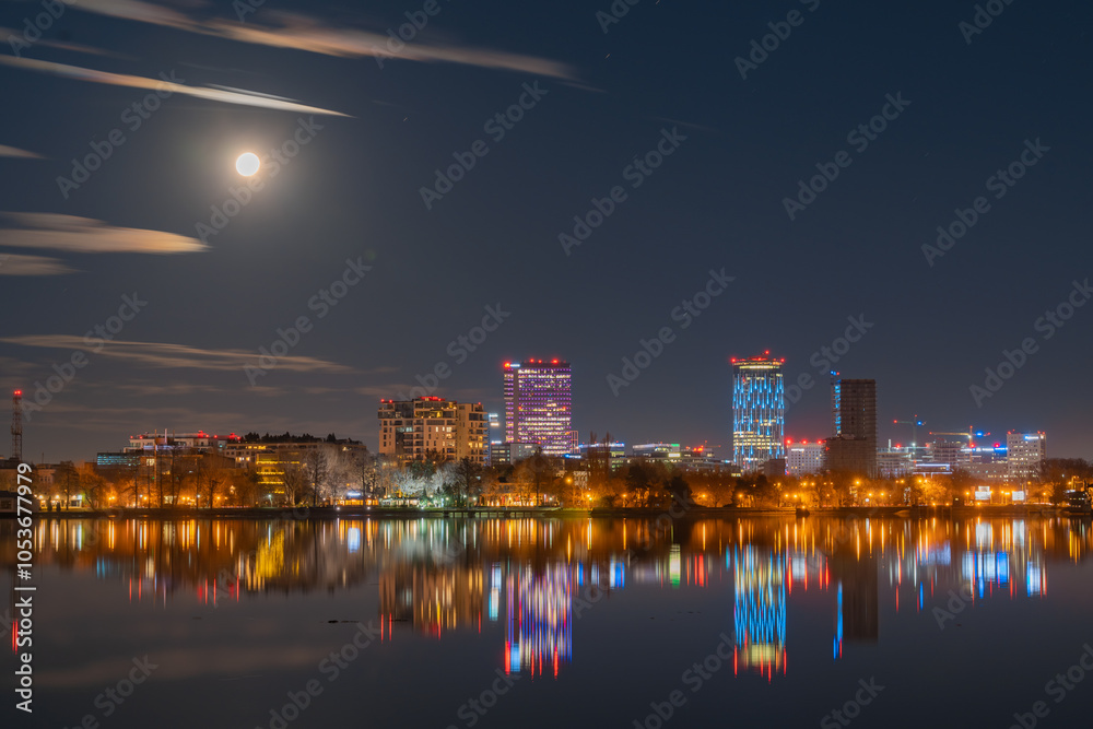 Amazing urban landscape with tall buildings illuminated at night and reflected in the water. Moonrise above the skyscrapers of Bucharest, the capital of Romania Amazing urban landscape with tall buildings illuminated at night and reflected in the water. Moonrise above the skyscrapers of Bucharest, the capital of Romania