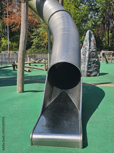 Grand toboggan tubulaire en métal, suspendu par des poteaux en bois, descend vers un sol en caoutchouc vert, avec des structures en bois en arrière-plan, aire de jeux pour enfant