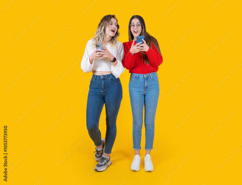 Fototapeta premium Shocked surprised happy two beautiful full body size front view young caucasian women using mobile phone looking touch screen isolated over yellow studio background. Wear casual jean and shirt.