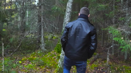 Man with black jacket and blue jeans walks past camera and disappears into the woods or forest ahead of him at day time day light. Fallen leaves are on the ground on this autumn cloudy day. photo