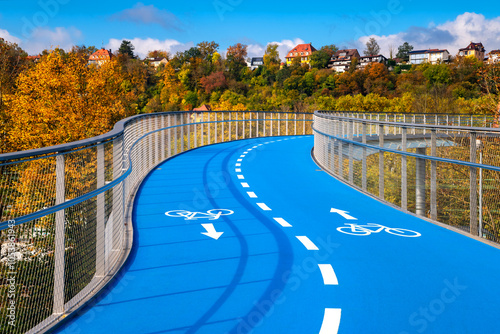 New built bicycle bridge in Tübingen (Germany) with curved, blue-colored roadway on a sunny autumn day. 2 cycle lanes with white lines and pictograms lead with incline in curves over a railroad. photo