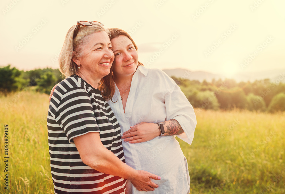 Obraz premium Portrait of a sincerely smiling young pregnant woman dressed in light summer clothes embracing with mother in evening sunset hours. Woman's health, human in the nature concept image.