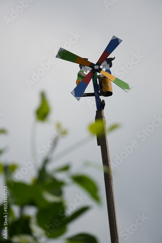 A colorful wooden windmill on a pole, producing a tin-like sound when spinning. Decorated with two cartoon figures whose arms move with each breeze-driven rotation.