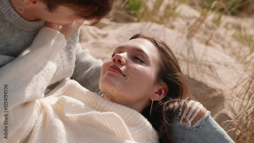 A young couple in love sharing a tender moment, with the woman lying on a man's lap, both dressed in cozy sweaters amidst a serene beach setting.
