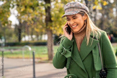 A cheerful woman enjoys a sunny autumn day in the park, staying connected through her smartphone, dressed in seasonal green outerwear and trendy accessories.