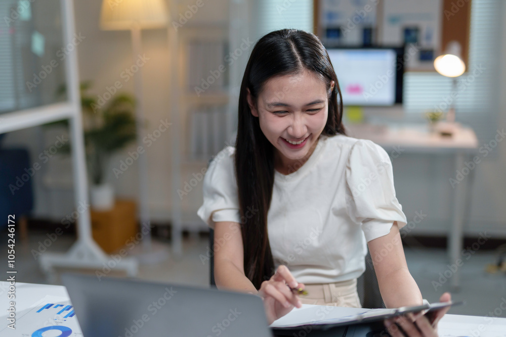Happy young asian businesswoman smiling and checking financial documents while working on laptop at her desk in modern office Happy young asian businesswoman smiling and checking financial documents while working on laptop at her desk in modern office
