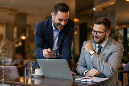 Принти на полотні Two mid-adult Caucasian businessmen collaborate using a laptop in a trendy cafe, engrossed in work while dressed in sharp suits