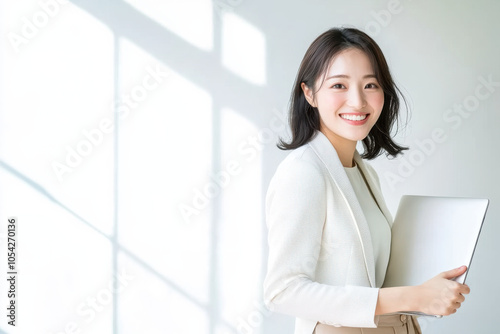 smiling woman in light colored suit holds laptop, standing in bright room with soft shadows. Her expression conveys confidence and professionalism photo