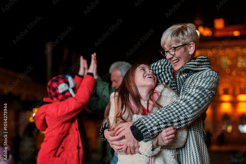 Fototapeta premium A joyful Caucasian senior woman embraces her young granddaughter, both smiling brightly, sharing a tender moment during an evening festive event, with blurred figures and lights in the background.