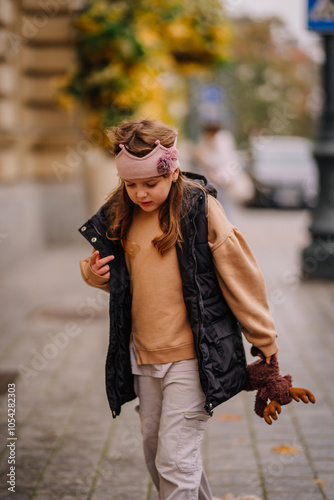 A young girl wearing a headband and vest walks down a street, holding a stuffed toy. Autumn foliage is visible in the background.. photo