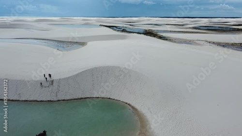 A drone reveals the vast white dunes of Lençóis Maranhenses, with shimmering lagoons nestled between the rolling sands. The sweeping aerial view captures the stunning contrast of the endless desert.