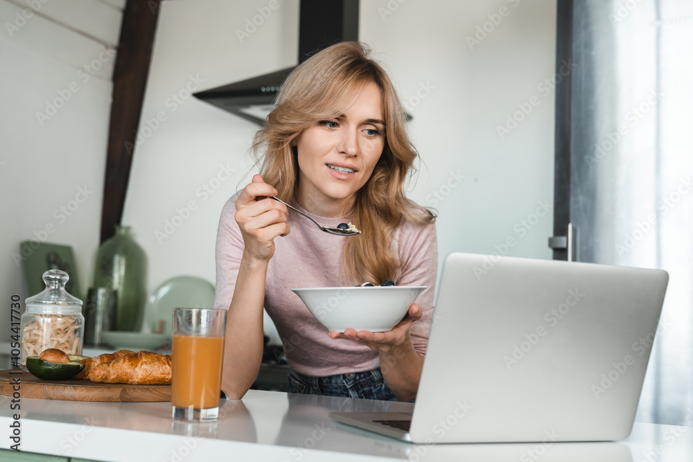 Attractive Caucasian woman with blond wavy hair casual outfit having healthy breakfast eating muesli with berries using laptop work remotely watching video reading news sitting in the cozy kitchen. Attractive Caucasian woman with blond wavy hair casual outfit having healthy breakfast eating muesli with berries using laptop work remotely watching video reading news sitting in the cozy kitchen.