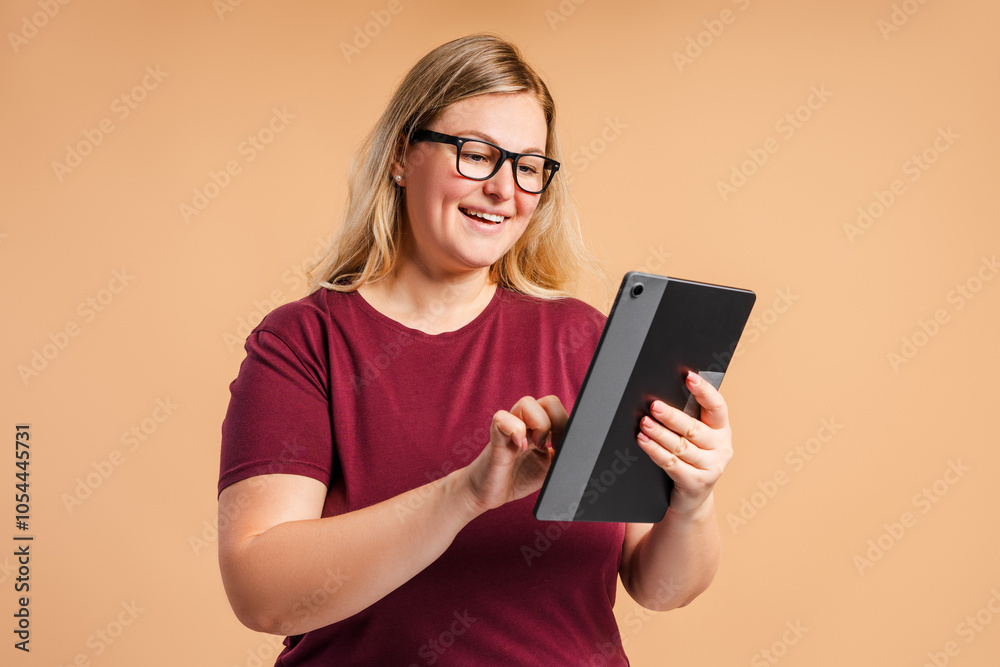 Smiling plus size woman in glasses typing on digital tablet isolated on beige background Smiling plus size woman in glasses typing on digital tablet isolated on beige background
