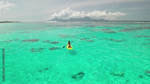 Woman is enjoying a sunny day on a yellow paddle board, surrounded by the turquoise water of a tropical lagoon with mountains in the background