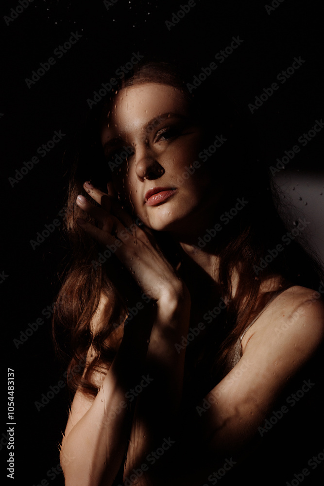 A woman looks at the camera through a window with raindrops visible on it. The woman is thoughtful and holds a green plant in her hands. A woman looks at the camera through a window with raindrops visible on it. The woman is thoughtful and holds a green plant in her hands.
